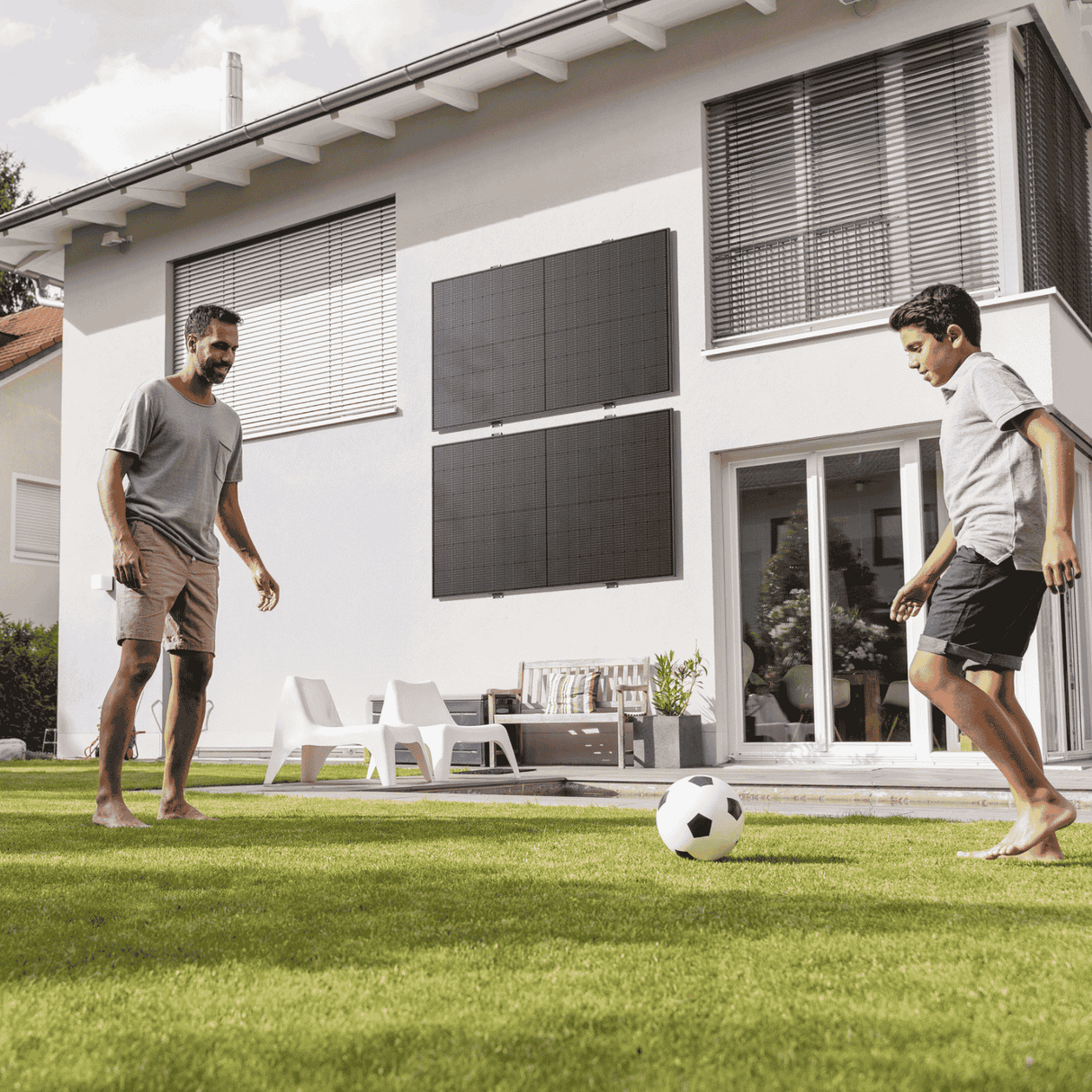 Balkonkraftwerk mit Solarmodulen an der Hausfassade eines Eigenheims mit zwei Personen beim Fußballspielen im Garten