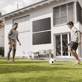 Balkonkraftwerk mit Solarmodulen an der Hausfassade eines Eigenheims mit zwei Personen beim Fußballspielen im Garten